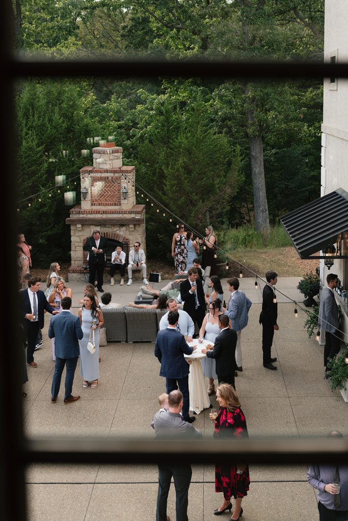 Outdoor wedding cocktail hour with guests mingling on patio near stone fireplace and string lights