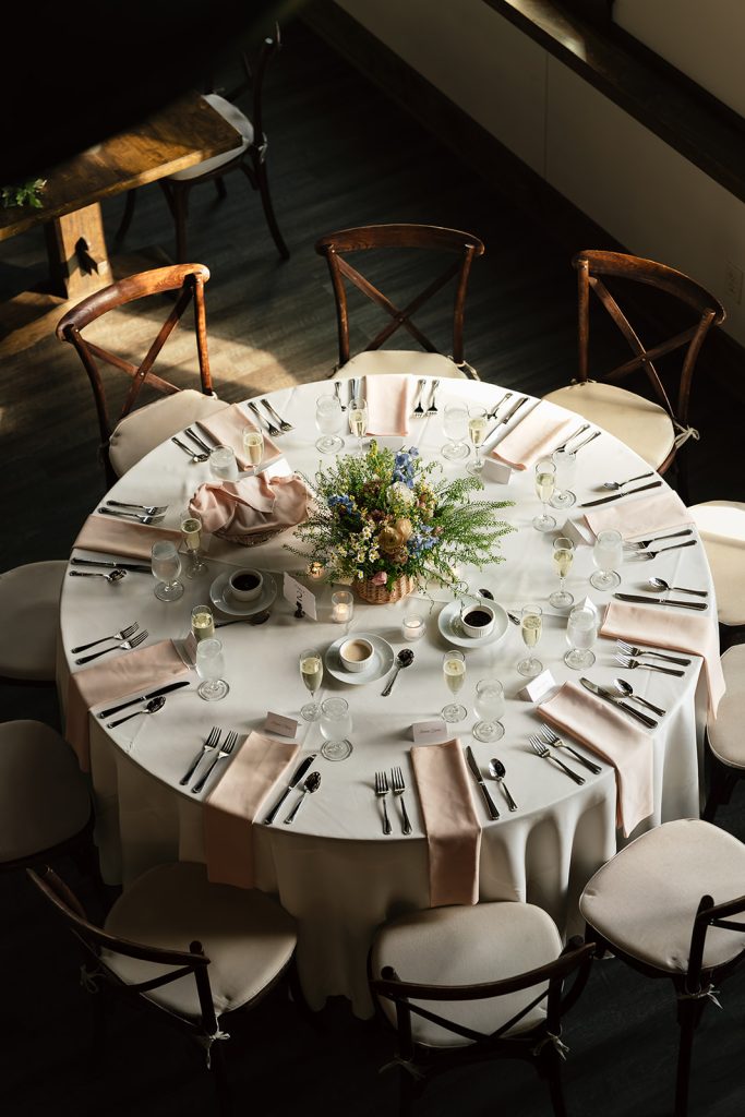 Round reception table with white linens, blush napkins, and greenery centerpiece bathed in natural light
