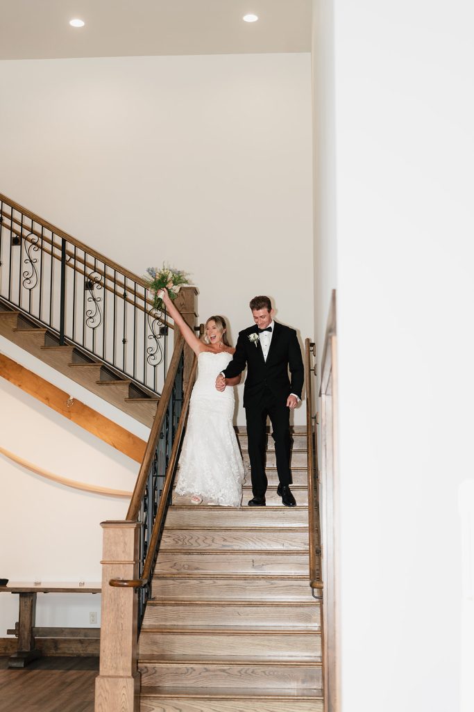 Newlyweds descending grand wooden staircase with wrought iron railing at reception venue