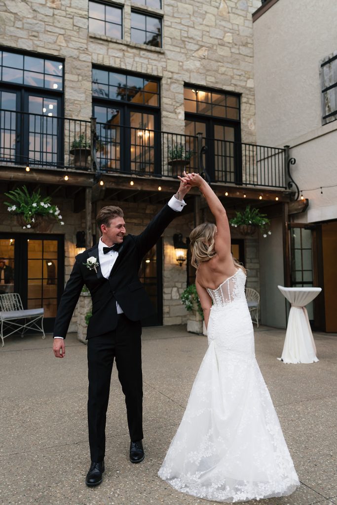 Groom twirling bride during first dance on courtyard patio at dusk under string lights