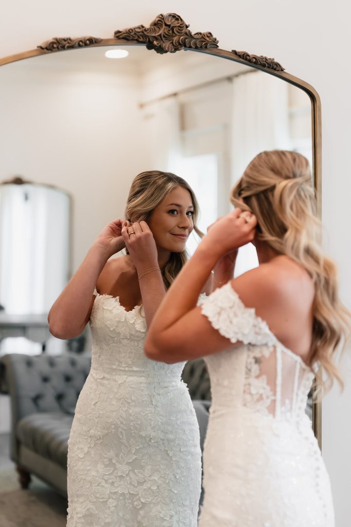 Bride Rachel putting on earrings while looking in ornate vintage mirror during wedding preparation