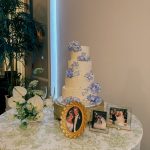 White tiered wedding cake displayed on table with white linen, alongside floral arrangements and framed photos