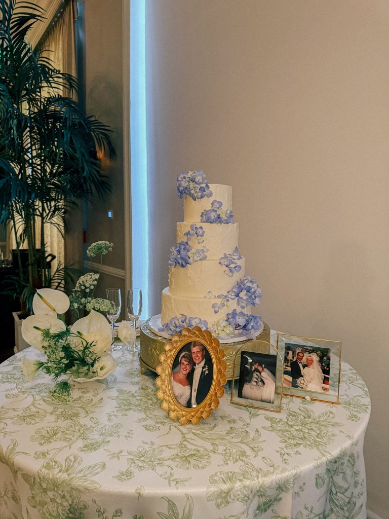 White tiered wedding cake displayed on table with white linen, alongside floral arrangements and framed photos