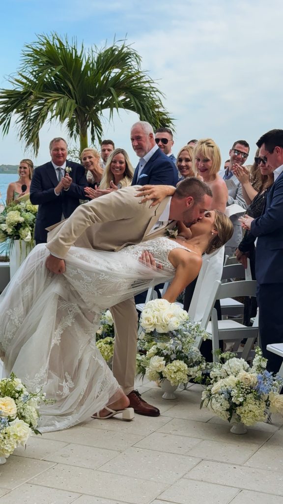 Groom dipping bride for dramatic kiss at waterfront ceremony as guests applaud under palm frond arch
