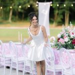 Bride in short white reception dress smiling at pink-themed outdoor wedding table with floral arrangements