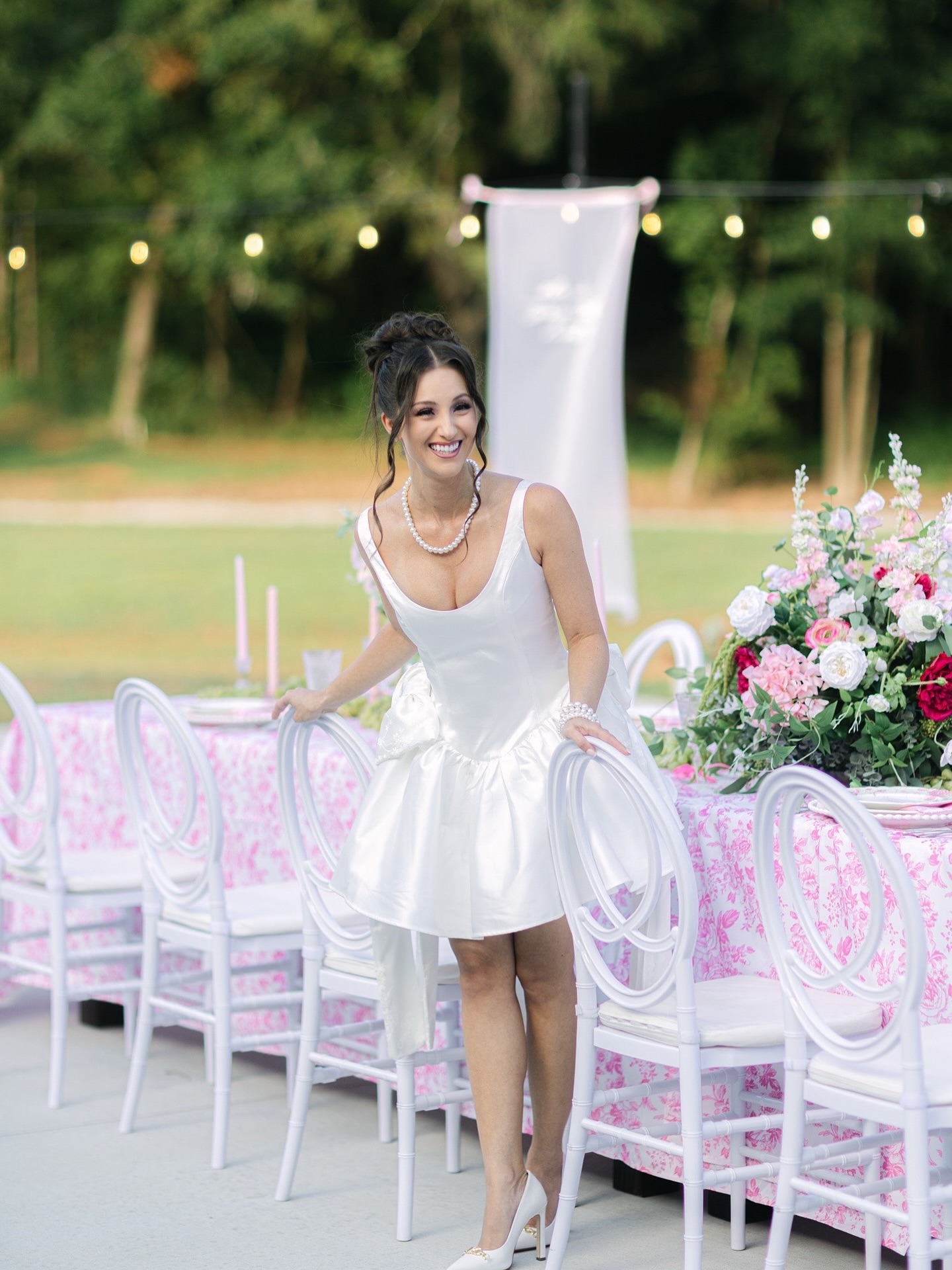 Bride in short white reception dress smiling at pink-themed outdoor wedding table with floral arrangements