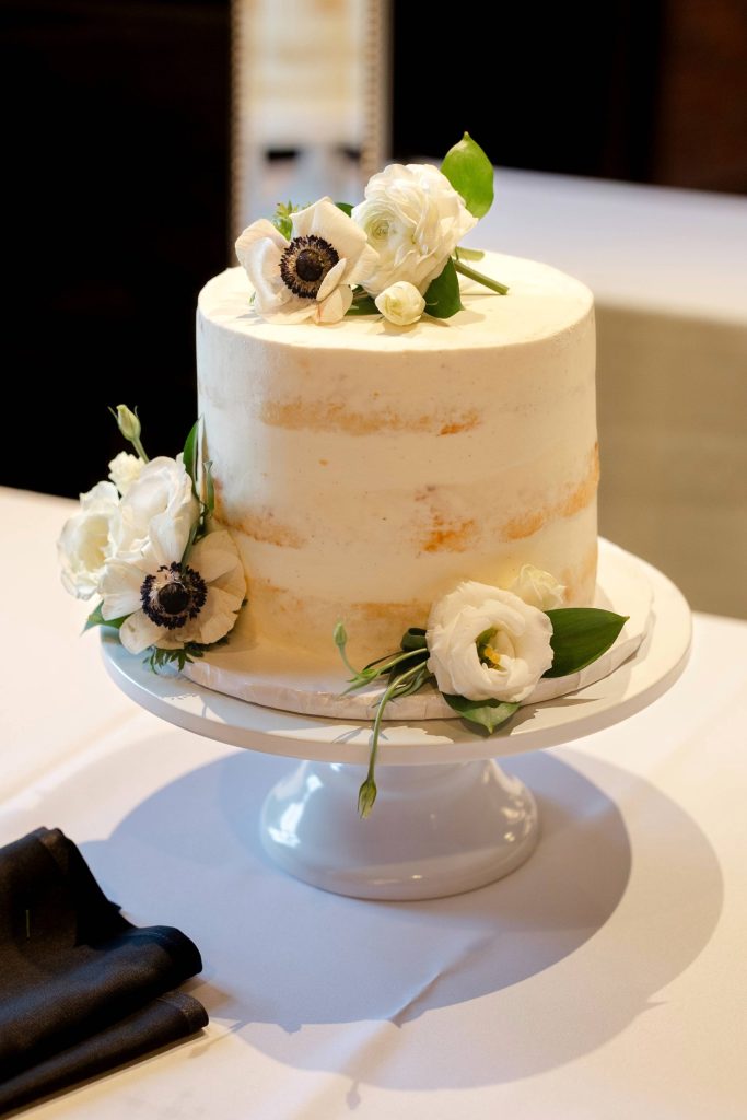 Two-tier semi-naked wedding cake with white buttercream on white cake stand, decorated with white anemones and ranunculus