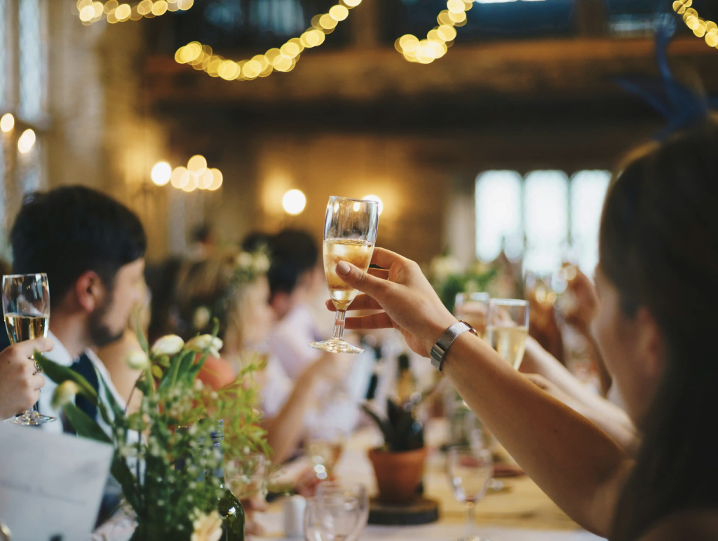 Wedding guests raising champagne glasses in toast at elegantly lit reception with string lights and floral centerpieces