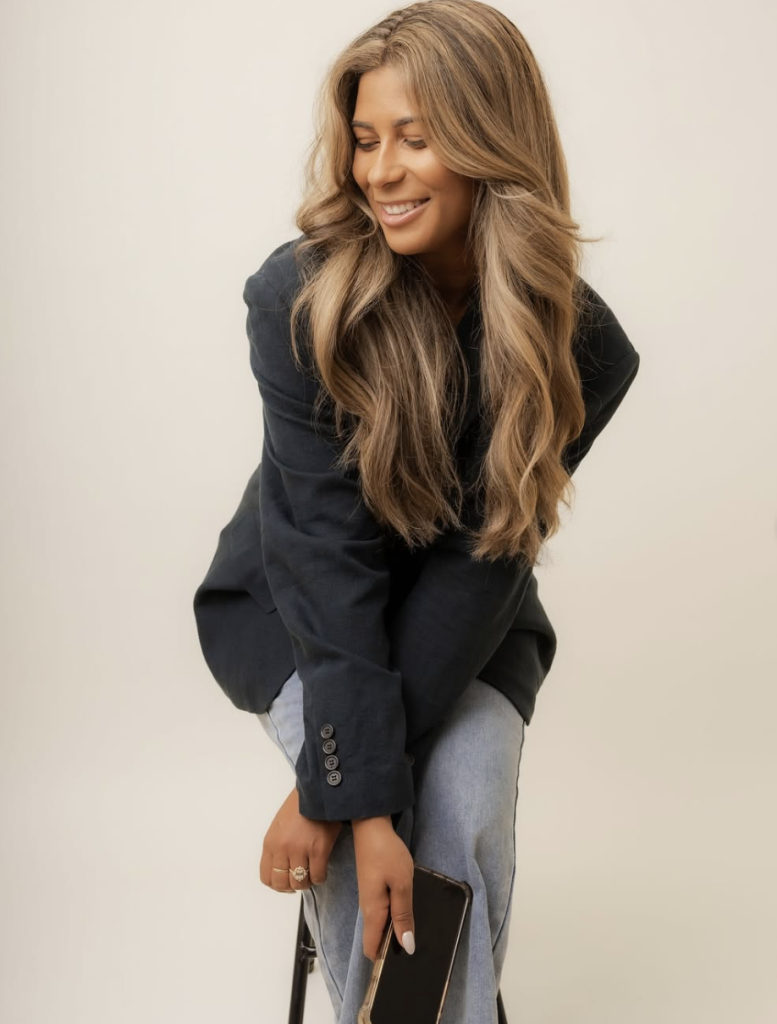 Professional woman in black blazer and gray pants smiling while holding smartphone against neutral background