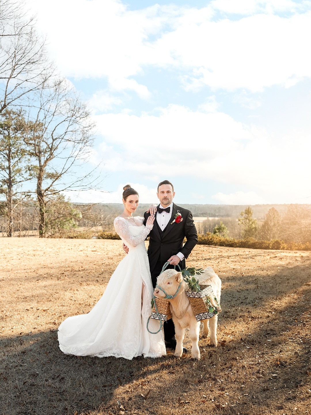 Bride and groom with decorated goat in rustic outdoor setting with rolling hills