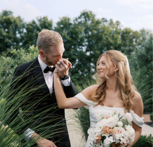 Groom in black tuxedo kissing bride's hand as she holds blush and white bouquet in outdoor garden setting