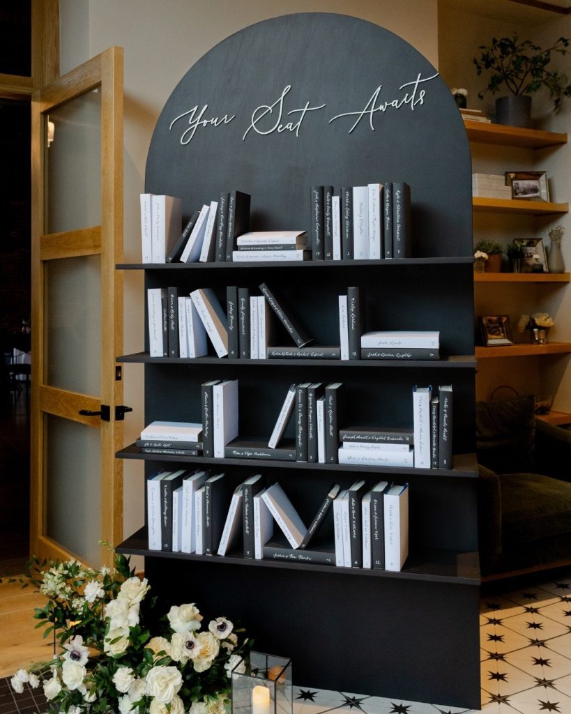 Black arched bookshelf seating chart display reading "Your Seat Awaits" with white floral arrangement at base