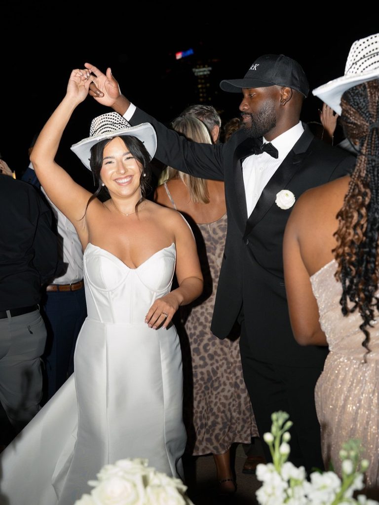 Katie and Anthony dancing at their rooftop reception, both wearing white cowboy hats with the St. Louis Gateway Arch illuminated in the background