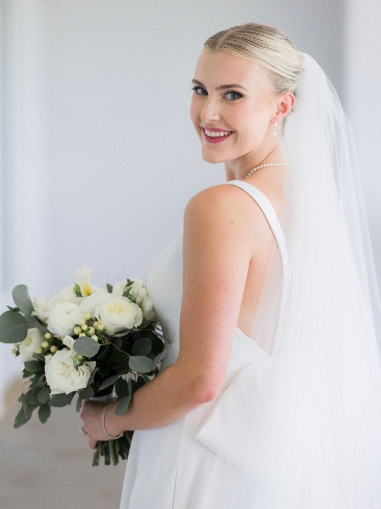 Bride Catherine smiling over shoulder holding white rose and eucalyptus bouquet in sleeveless gown with veil