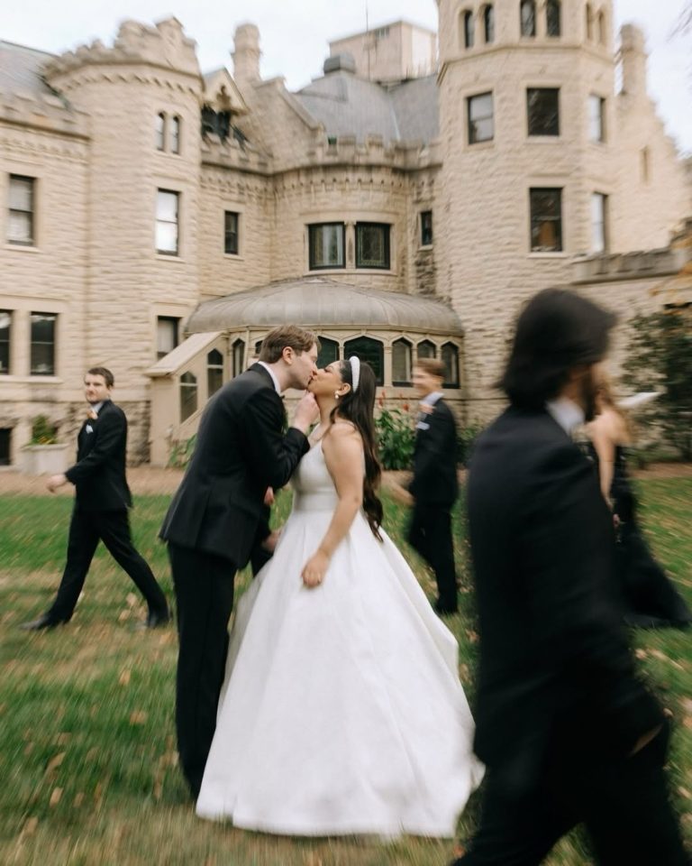 Newlyweds kissing outside historic stone building with wedding party walking on lawn