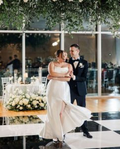 Bride and groom embracing under greenery installation at modern wedding venue with white floral arrangements