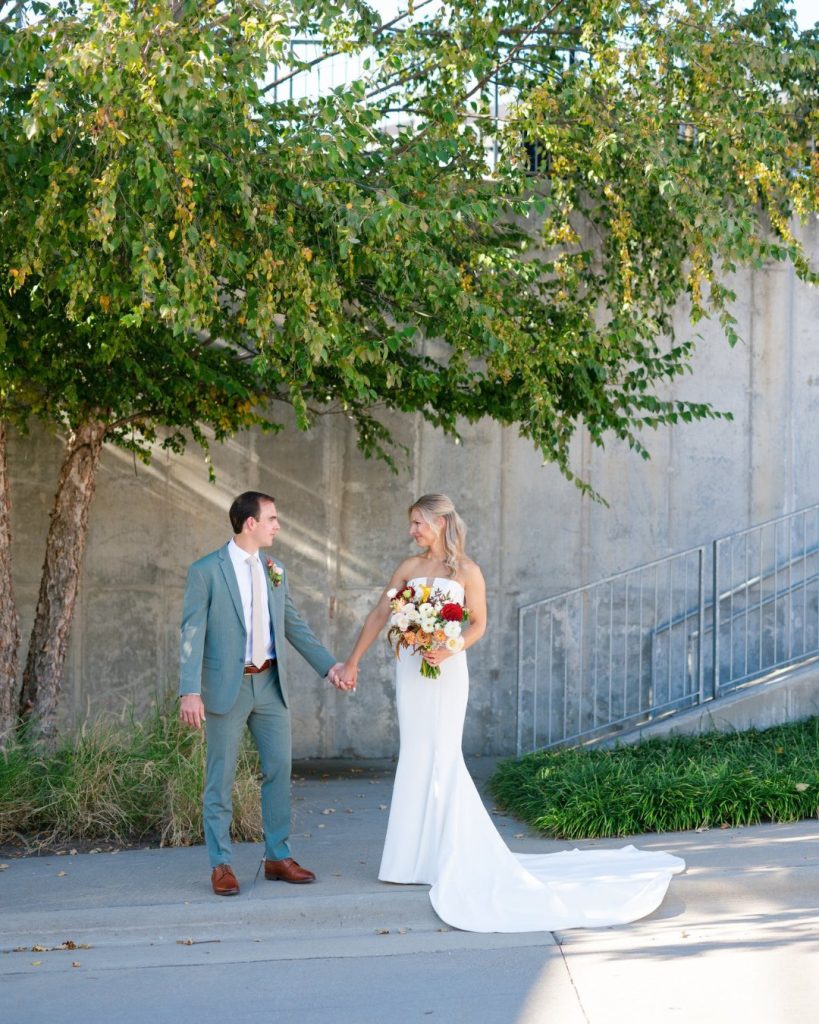 Bride and groom holding hands under leafy tree canopy against modern concrete wall in urban downtown setting