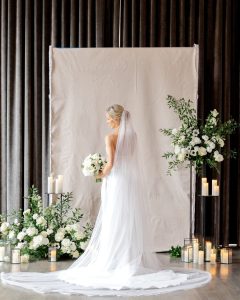 Bride in flowing white gown with cathedral veil holding white bouquet, framed by pillar candles and white floral arrangements
