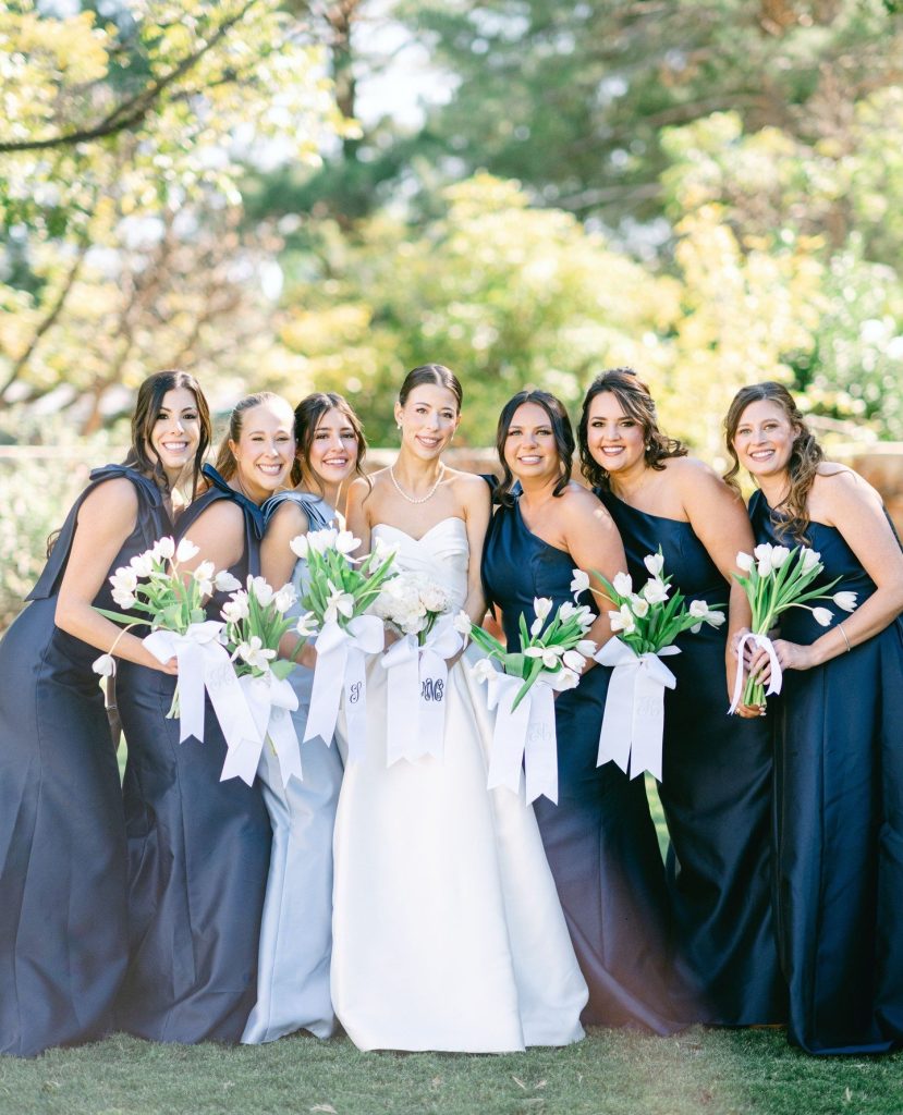 Bride in white gown with bridesmaids in navy dresses holding white tulip bouquets in outdoor garden setting