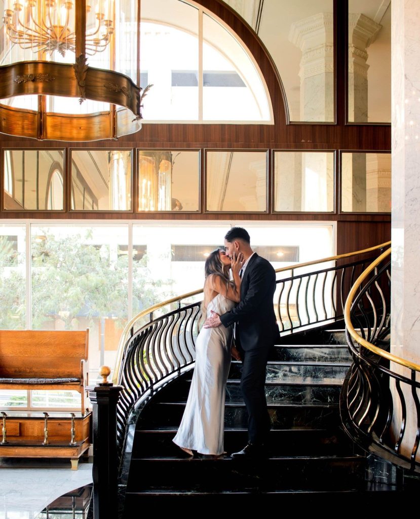 Bride and groom kissing on grand staircase at Hotel Paso Del Norte with ornate chandeliers and arched windows