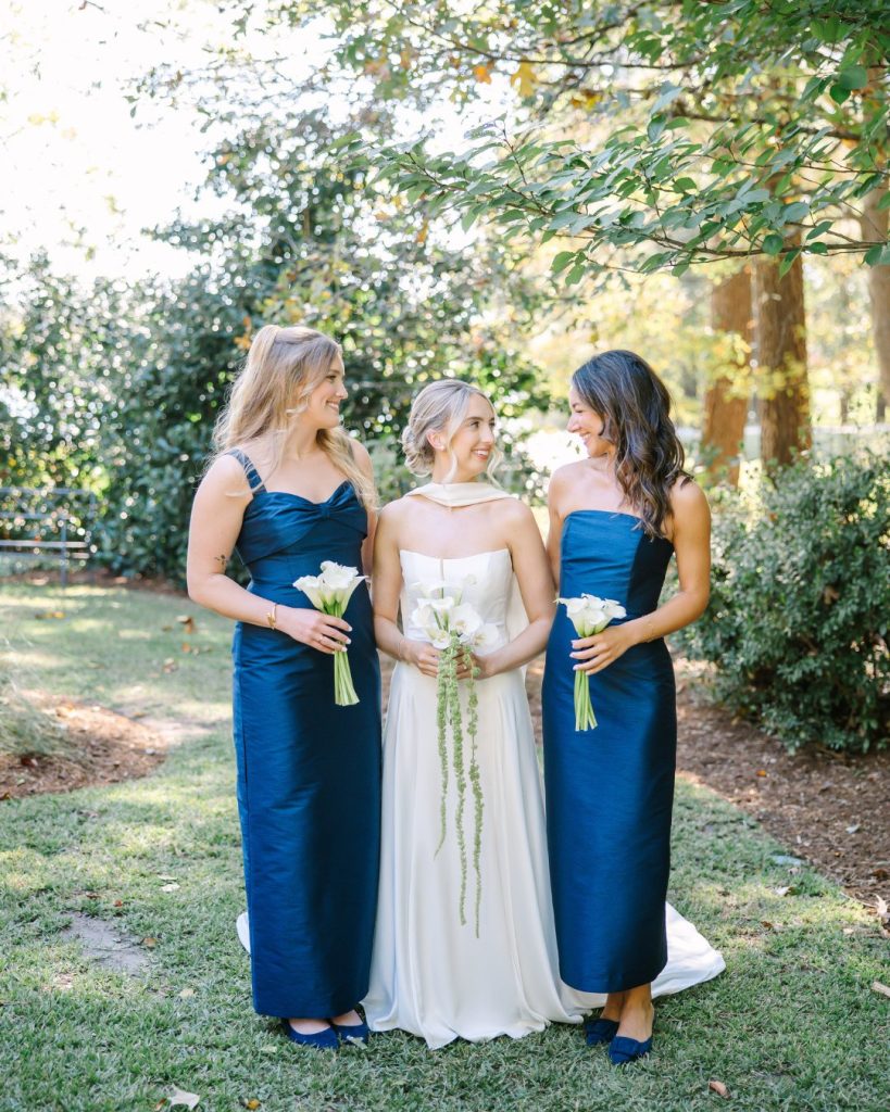 Bride in cream gown with two bridesmaids in navy blue dresses holding white calla lily bouquets in garden setting