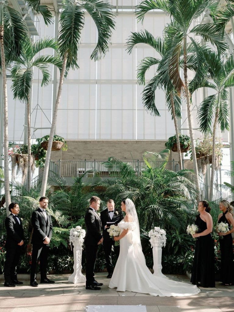 McKenzi and Alex exchange vows under tall palm trees in an airy conservatory with their black-attired wedding party