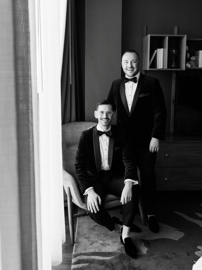 Stephen and Austin pose in matching black tuxedos with bow ties in an elegant indoor setting before their wedding ceremony