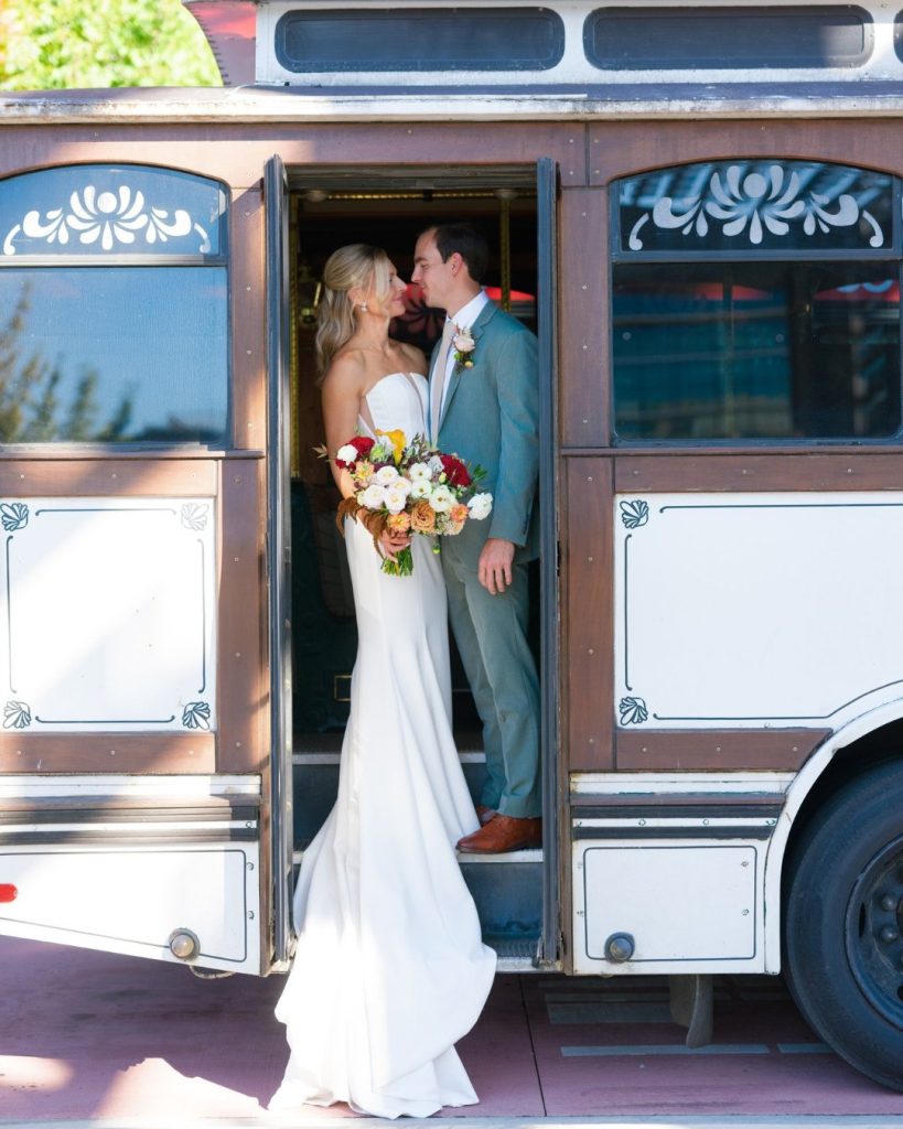 Newlyweds kissing in doorway of vintage trolley, bride holding colorful bouquet against her strapless white gown