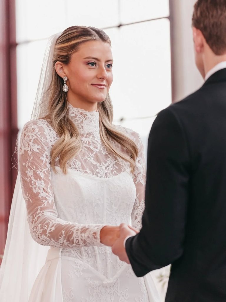 Bride in lace wedding gown with veil holding hands with groom in black suit during ceremony