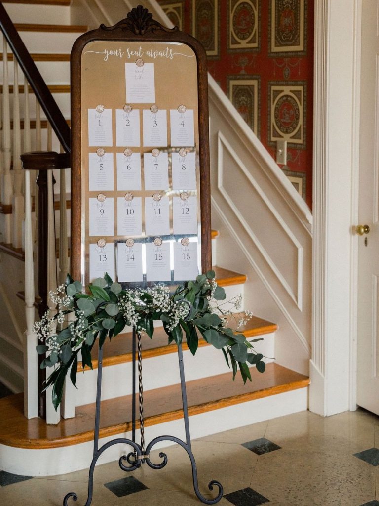 Vintage mirror seating chart displaying numbered table assignments adorned with eucalyptus and baby's breath garland