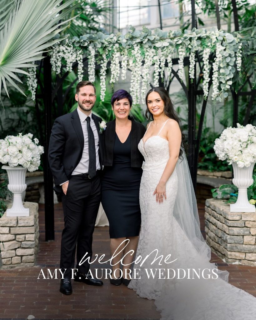 Bride and groom pose with wedding planner beneath white floral arch in conservatory setting