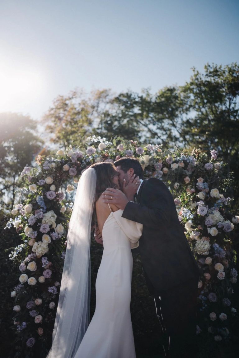 Couple sharing first kiss under romantic circular floral arch filled with blush roses and lavender blooms