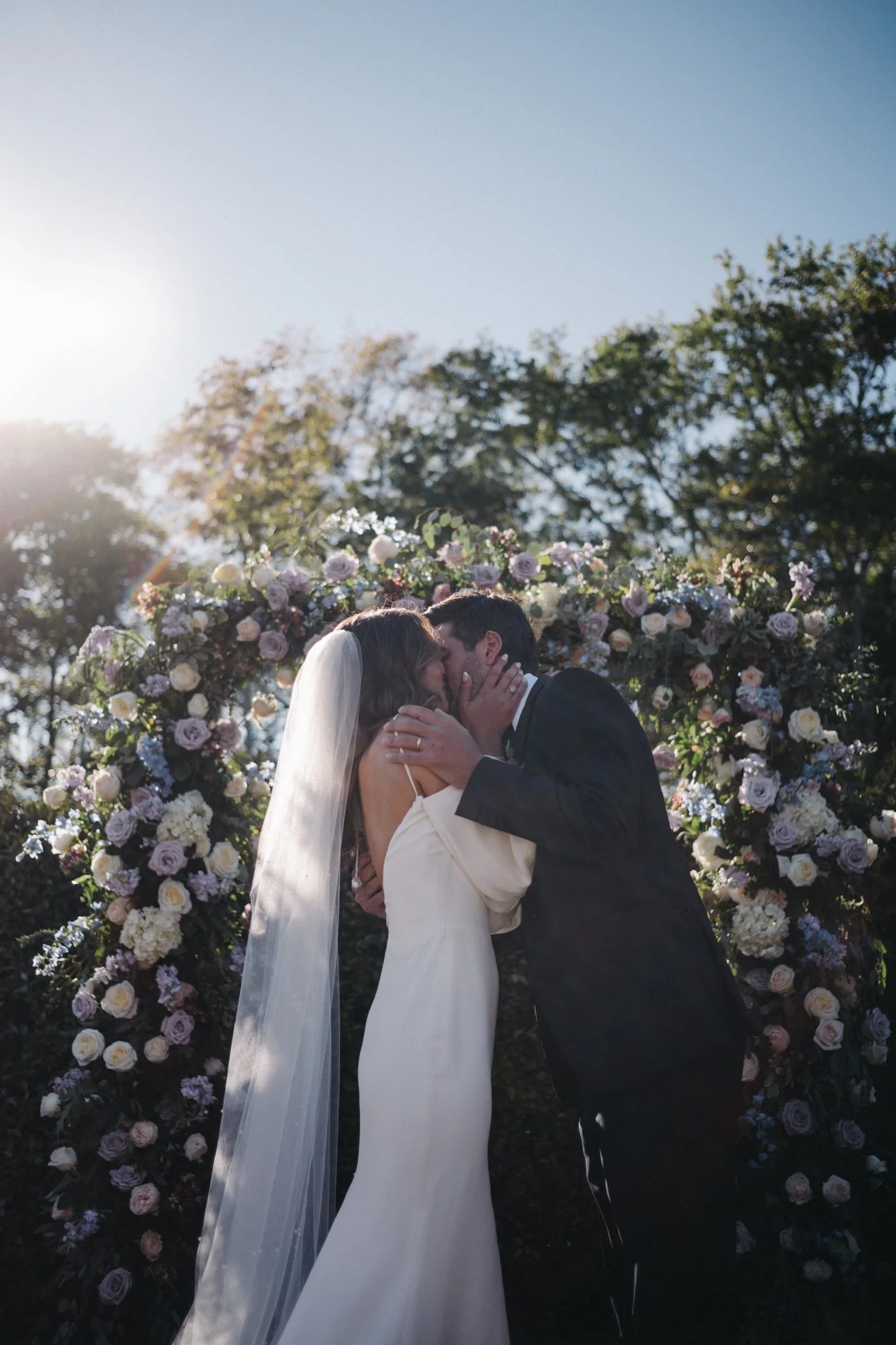 Couple sharing first kiss under romantic circular floral arch filled with blush roses and lavender blooms