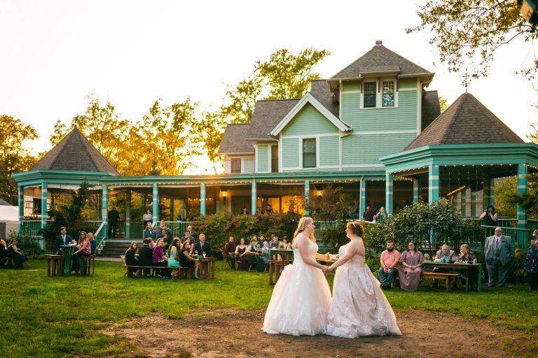 Two brides holding hands during outdoor ceremony at Victorian house at golden hour