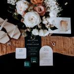 Wedding flat lay with pearl-embellished sandals, rose bouquet, invitation suite, and emerald ring box on rust-colored fabric