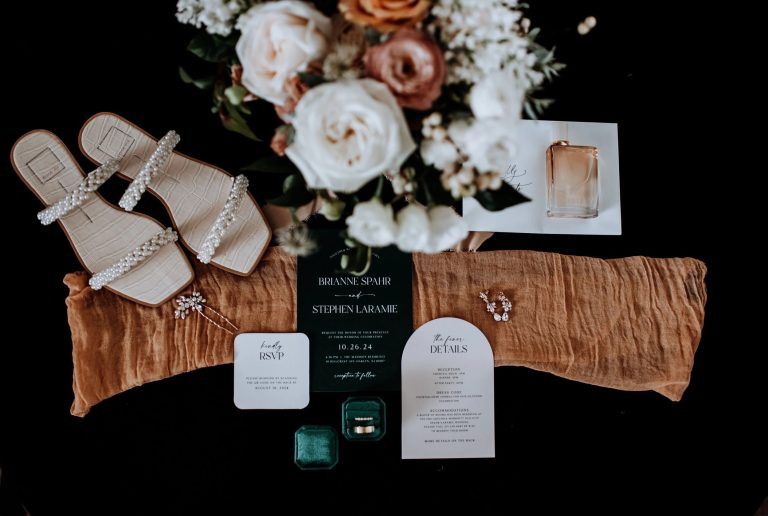 Wedding flat lay with pearl-embellished sandals, rose bouquet, invitation suite, and emerald ring box on rust-colored fabric