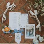 Flat lay of wedding stationery suite with ribbons and floral accents on terracotta surface