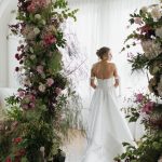 Bride in off-shoulder white gown standing beneath floral arch with pink, burgundy, and green blooms in bright indoor venue