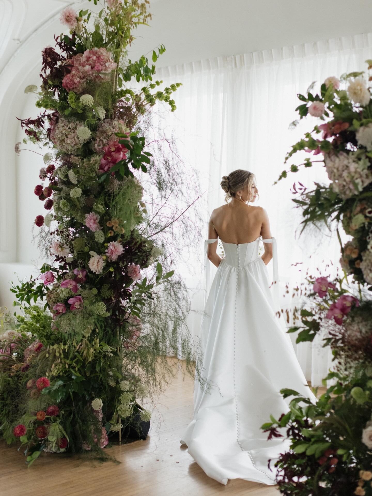 Bride in off-shoulder white gown standing beneath floral arch with pink, burgundy, and green blooms in bright indoor venue