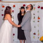 Groom laughing during wedding ceremony while bride smiles and officiant holds microphone, framed by colorful dahlias on white backdrop