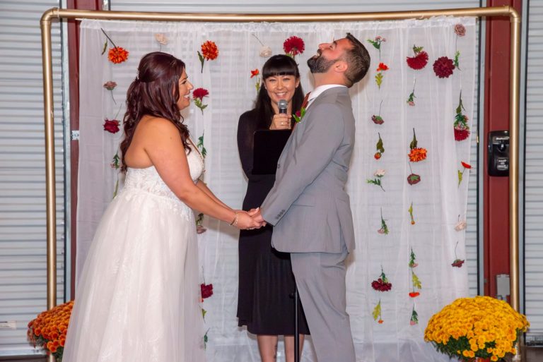 Groom laughing during wedding ceremony while bride smiles and officiant holds microphone, framed by colorful dahlias on white backdrop