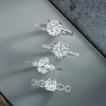 Four diamond engagement rings in varied styles displayed on gray surface, featuring solitaire, halo, and three-stone designs