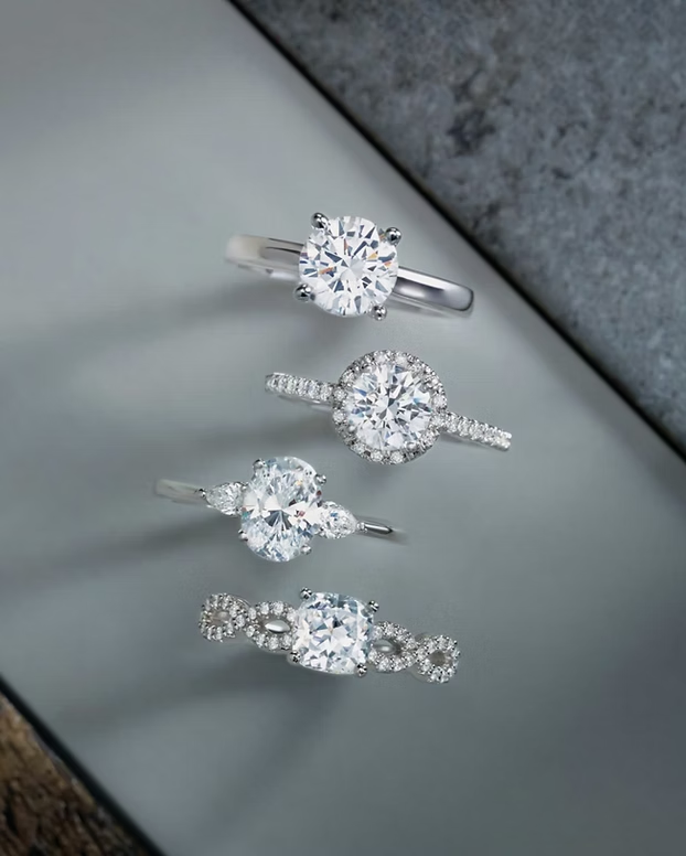Four diamond engagement rings in varied styles displayed on gray surface, featuring solitaire, halo, and three-stone designs