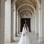 Bride in long-sleeved lace gown standing in stone corridor framed by elegant archways