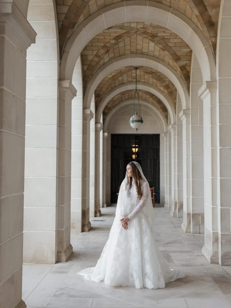 Bride in long-sleeved lace gown standing in stone corridor framed by elegant archways