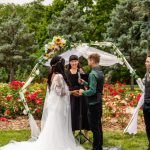 Wedding officiant conducting outdoor ceremony under floral arch with couple exchanging vows in rose garden