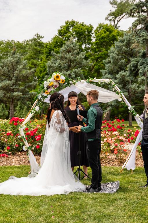 Wedding officiant conducting outdoor ceremony under floral arch with couple exchanging vows in rose garden