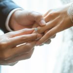Groom placing diamond engagement ring on bride's finger during wedding ceremony