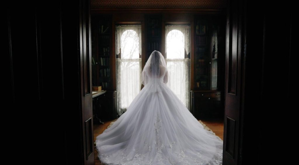 Bride in voluminous ballgown and veil standing before bright arched windows in dark vintage room