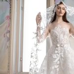 Bride wearing sheer lace wedding gown with elaborate floral appliqués and oversized statement cape sleeves